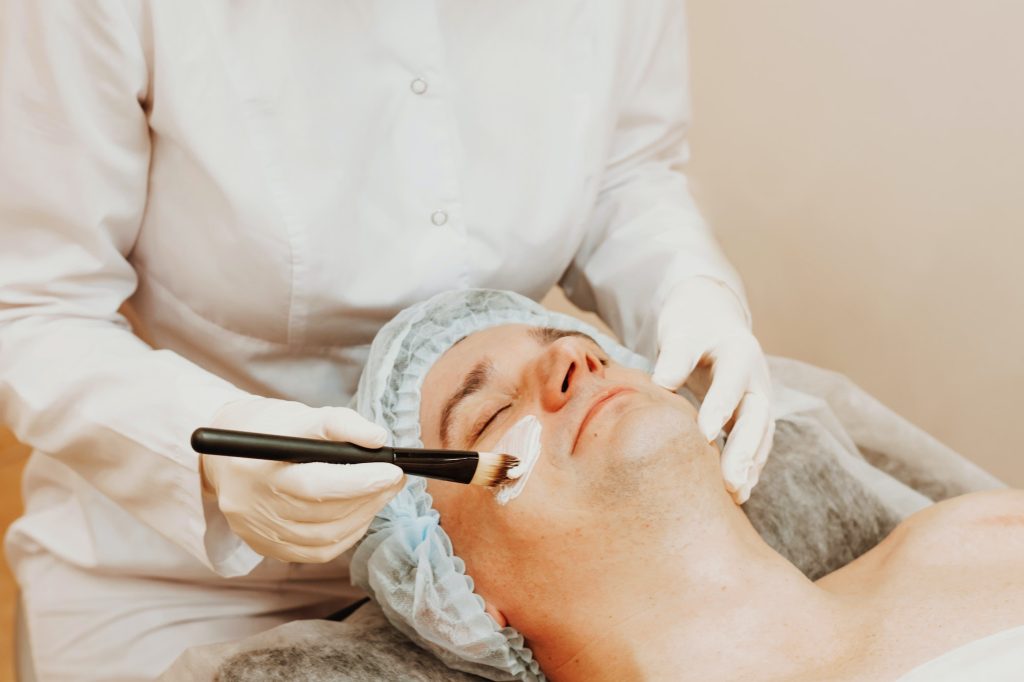 Beautician applying scrub mask on face to young man, spa beauty salon. The concept of body and face